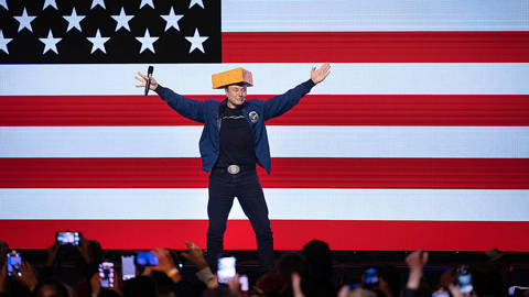 Elon Musk wearing a cheese hat in front of US flag.
