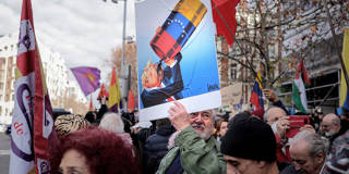 A protester in Madrid holds up a caricature by Portuguese cartoonist Vasco Gargalo depicting U.S. President Donald Trump eagerly drinking from a Venezuelan oil barrel, during a demonstration on January 4, 2026, against the U.S. military operation in Venezuela that resulted in the capture of Venezuelan President Nicolás Maduro.