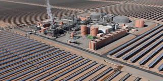 Aerial view of the Noor 1 concentrated solar power plant in Morocco, showing a vast field of solar mirrors arranged in a circular and grid-pattern reflecting sunlight in a desert landscape.