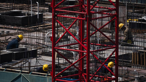 Construction workers in yellow helmets work on a reinforced concrete structure at a Chongqing building site, with a red crane in the foreground, illustrating China’s infrastructure investment and economic stimulus efforts.