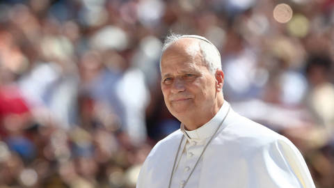 Pope Leo XIV stands in St. Peter’s Square during a general audience at the Vatican, wearing white papal vestments and a white skullcap. He smiles and raises one hand in greeting while speaking to a large crowd gathered behind barriers.