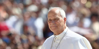 Pope Leo XIV stands in St. Peter’s Square during a general audience at the Vatican, wearing white papal vestments and a white skullcap. He smiles and raises one hand in greeting while speaking to a large crowd gathered behind barriers.