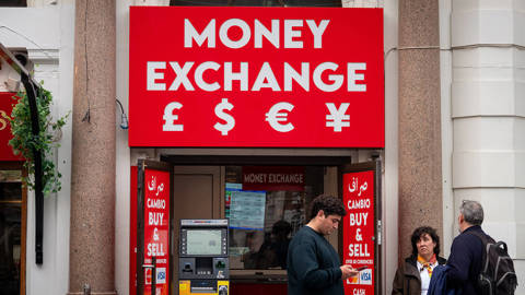 People stand outside a currency exchange displaying exchange rates for major currencies, including the US dollar, euro, and British pound.