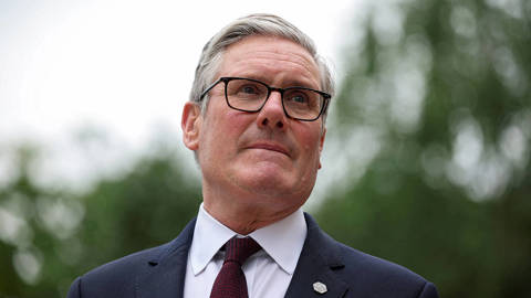 British Prime Minister Keir Starmer standing in a formal government setting, looking ahead with a serious expression during an official engagement, with aides and institutional surroundings in the background.