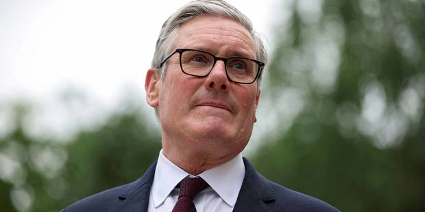 British Prime Minister Keir Starmer standing in a formal government setting, looking ahead with a serious expression during an official engagement, with aides and institutional surroundings in the background.
