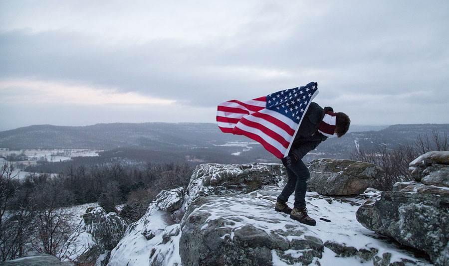 Man holding an American flag in snowy mountains.